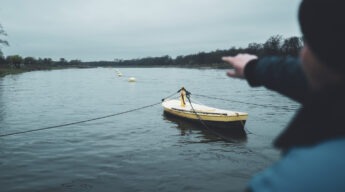 Ein Fährmann zeigt auf die gelben Bojen der Gierfähre Coswig (Anhalt), die über die Elbe gespannt sind. Im Vordergrund liegt das gelbe Steuerboot, der Himmel ist grau und die Stimmung ruhig.
