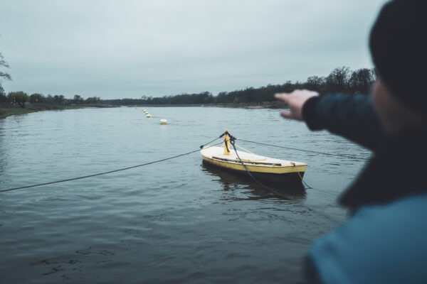 Der Fährmann 1 Ein Fährmann zeigt auf die gelben Bojen der Gierfähre Coswig (Anhalt), die über die Elbe gespannt sind. Im Vordergrund liegt das gelbe Steuerboot, der Himmel ist grau und die Stimmung ruhig.